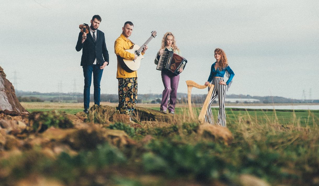 The image shows four band members stood on a grass hill, each with an instrument.