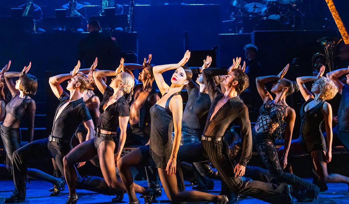 Image shows a stage in blue lighting, there are a number of dancers dressed in all black on one knee leaning backwards with their hand on their heads.