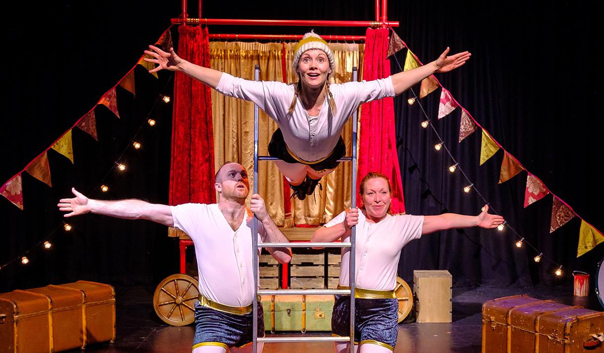 The image shows three people on stage, two men are holding a ladder from either side at the bottom of it. There is a woman with her waist on the ladde