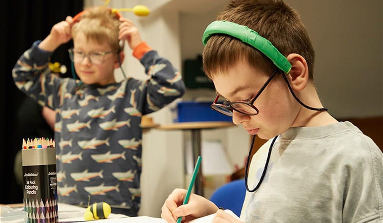 The image shows two young children, one is stood up whilst the other is sat down at a table drawing on a piece of paper.