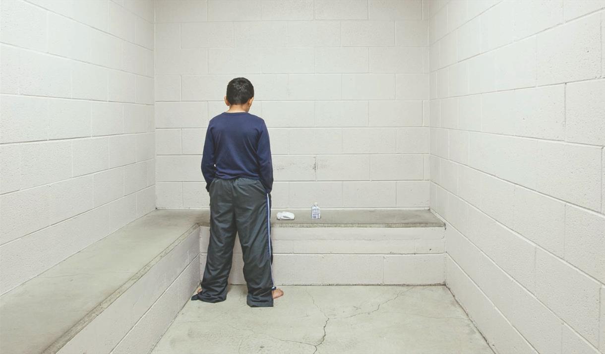 Photo from the exhibition showing a young person chained, facing a blank wall
