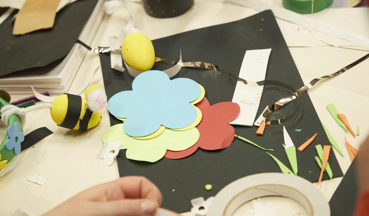 Image shows a craft table with coloured pieces of paper on it in the shape of flowers and a hand pulling some tape off a roll.