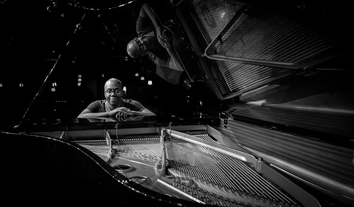 Black and white image of a man stood at a piano, the piano is open and the inside pipes can be seen.