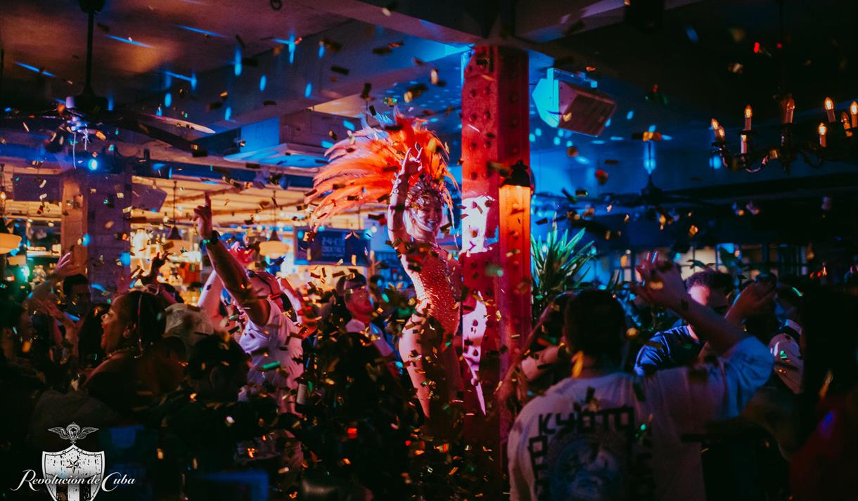 Photo of a crowded bar with fantastic dancing performers in feather headpieces, glitter faling from the ceiling and disco lights.