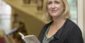 A woman with shoulder length blonde hair stands in a library holding a book